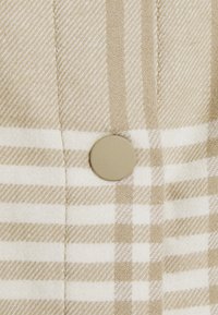 Tissu à carreaux beige et blanc avec des rayures diagonales et verticales, avec un grand bouton beige mat cousu près du centre.