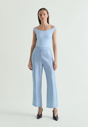 Vrouw draagt een lichtblauw off-shoulder topje en bijpassende wijde broek, staat voor een effen lichte achtergrond, draagt zwarte puntige hakken.