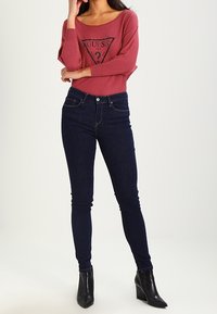 Top rosa, con scollatura a spalle scoperte e maniche lunghe, decorato con un logo grafico nero; abbinato a jeans attillati blu scuro e stivaletti neri alla caviglia.
