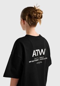 Osoba z brązowymi włosami związanymi w koka, ubrana w czarny, oversize'owy t-shirt z białym napisem "ATW OFF DUTY GEAR HALO" na plecach.