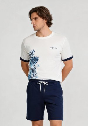 Młody mężczyzna w białej koszulce z niebieskim kwiatowym wzorem i napisem "California", w połączeniu z granatowymi szortami z białym sznurkiem.