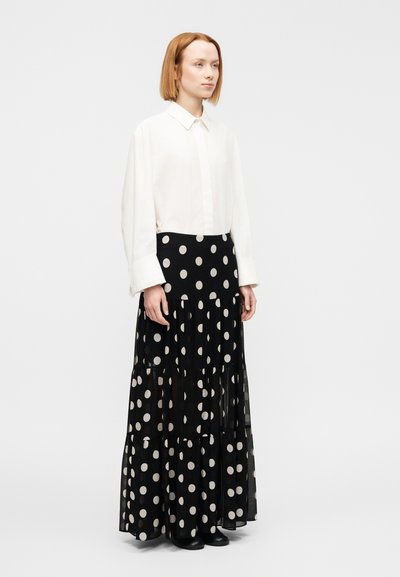 Λευκό μακρυμάνικο πουκάμισο με γιακά, συνδυασμένο με μαύρη φούστα μ ε βολάν και μεγάλα μπεζ πουά. Οι μπότες είναι μαύρες και το φόντο απλό.