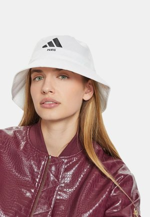 Donna con capelli biondi lunghi indossa un cappello a secchiello bianco Adidas Paris e una giacca con zip di colore amaranto lucido e struttura texturizzata su sfondo chiaro uniforme.