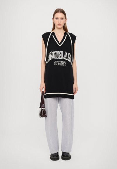 Túnica preta sem mangas em malha com detalhes brancos, apresentando um decote em V e estampa de logótipo. Combinada com calças largas de riscas e sapatos pretos robustos.