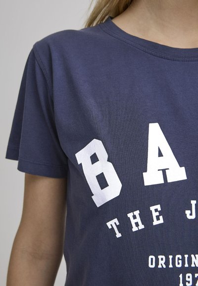 Tumedate sinine puuvillane T-särk lühikeste varrukatega, millel on julged valged tekstid: "BA", "THE J" ja "ORIGINAL 1973", pehme tekstuuriga.