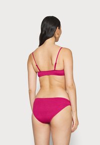 Magenta bikini komplektas, pasižymintis tekstūrinėmis medžiagomis, plonomis reguliuojamomis petnešėlėmis ir atitinkančiomis kelnaitėmis su vidutinio aukščio uždengimu.