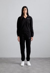 Zwarte zip-up hoodie met een voorzak, gecombineerd met bijpassende zwarte sweatpants. Witte sneakers maken de outfit af. Glad textuur en aansluitend ontwerp.