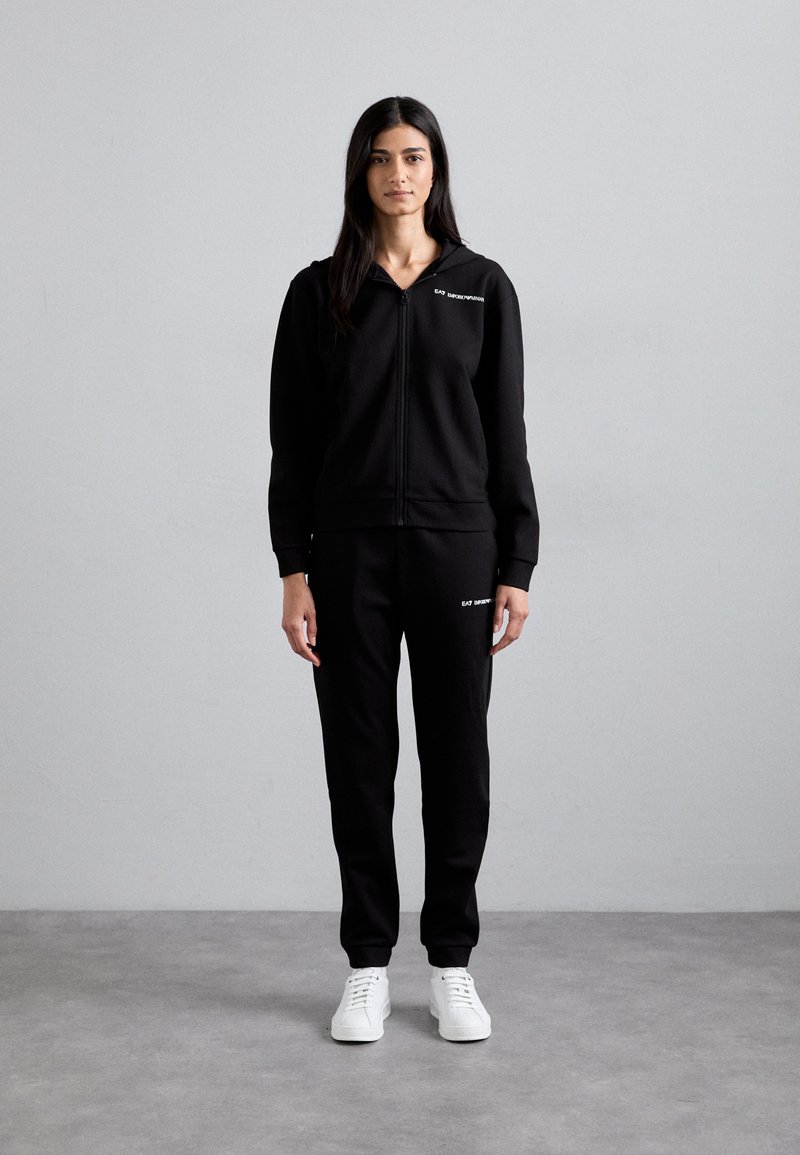 Zwarte zip-up hoodie met een voorzak, gecombineerd met bijpassende zwarte sweatpants. Witte sneakers maken de outfit af. Glad textuur en aansluitend ontwerp.