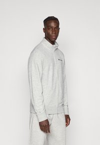 Ανοιχτό γκρι φούτερ με φερμουάρ, ψηλό γιακά και ριμπ μανσέτες. Διαθέτει κεντημένο λογότυπο στο στήθος. Μαλακή υφή φλις.