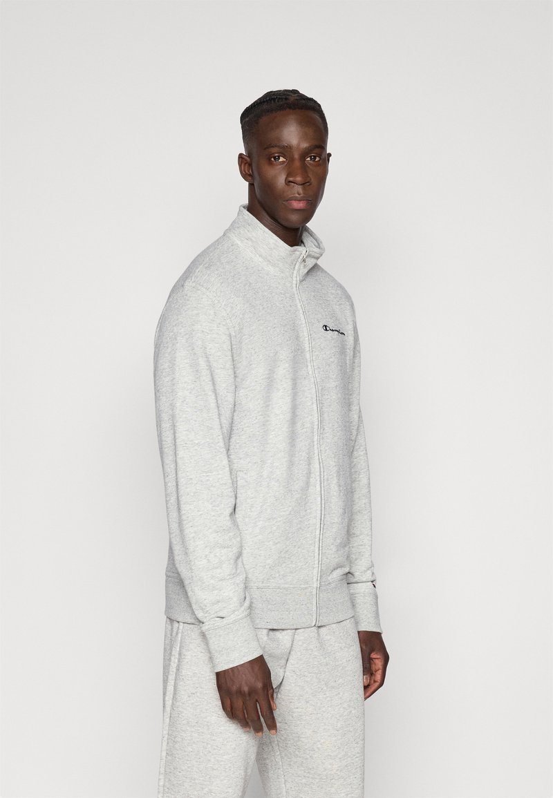 Ανοιχτό γκρι φούτερ με φερμουάρ, ψηλό γιακά και ριμπ μανσέτες. Διαθέτει κεντημένο λογότυπο στο στήθος. Μαλακή υφή φλις.