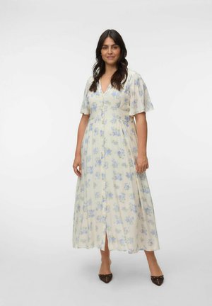 Frau steht, trägt ein hellcremefarbenes Maxikleid mit kurzen Ärmeln und blauem Blumenmuster, braune spitze Schuhe, neutraler Hintergrund.
