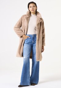 Abrigo de piel sintética beige con cuello de muesca, cierre de botones y mangas largas, combinado con jeans acampanados de color azul claro y zapatos de estampado animal.