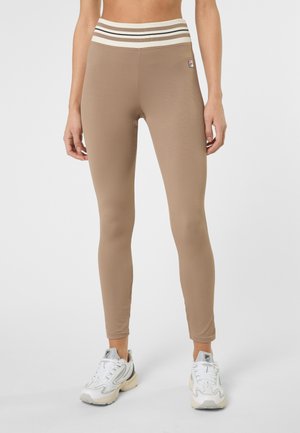 Kobieta ubrana w beżowe ściągane legginsy z wysokim stanem i paskiem w paski oraz białe sportowe buty, stojąca na jednolitym tle.