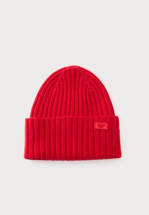 Gorro de punto rojo con canalé, con puño doblado y una pequeña etiqueta con logo de tela en el puño, sobre un fondo liso y claro.