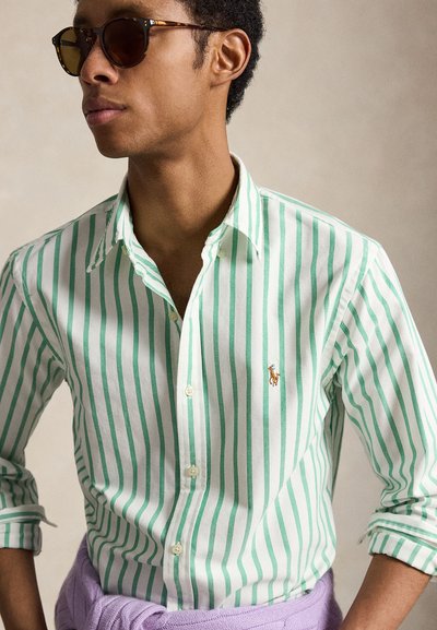 Άνδρας που φορά καφέ γυαλιά ηλίου και άσπρο πουκάμισο με πράσινες κάθετες ρίγες και μικρό κεντημένο λογότυπο στο στήθος.