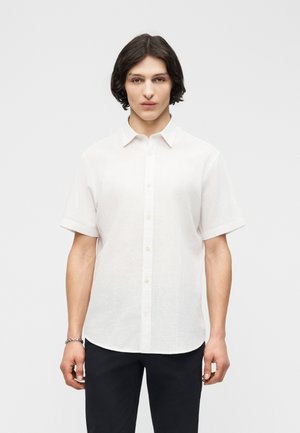 Jeune personne aux cheveux mi-longs foncés portant une chemise blanche à manches courtes boutonnée et un pantalon noir, sur un fond uni.