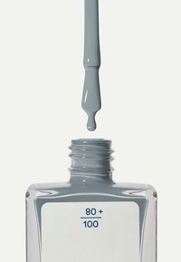 Esmalte de uñas gris en una botella de cristal transparente, con un cuello redondo. Una gota de esmalte cuelga del pincel, lista para ser aplicada.