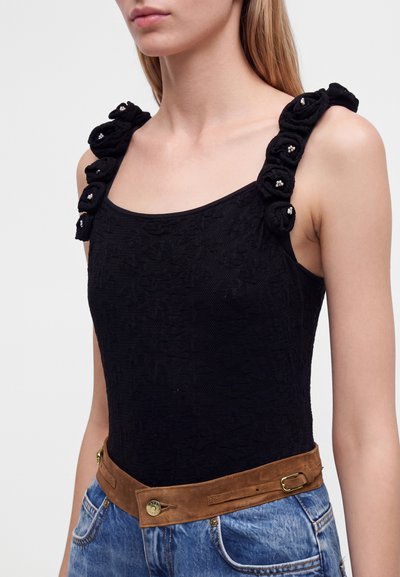 Γυναίκα που φοράει μαύρο αμάνικο τοπ με υφασμάτινη υφή και λουλουδάτες τιράντες, συνδυασμένο με μπλε τζιν και καφέ ζώνη.
