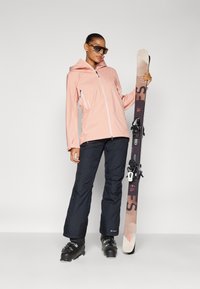 Růžová lyžařská bunda se zipy, černé lyžařské kalhoty, černé lyžařské boty. Model drží lyži s béžovo-hnědým designem. Hladké textury, funkční design.