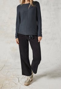Femme portant un haut texturé à manches longues de couleur foncée, un pantalon large en velours côtelé noir avec un cordon de serrage, des chaussettes grises et des baskets beiges, debout à l'intérieur.