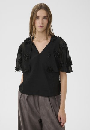 Frau trägt eine schwarze transparente Bluse mit floralen Applikationen an Ärmeln und Schultern, kombiniert mit lockeren braunen Nadelstreifenhosen.