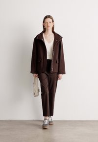 Brown zip-up jacket with a wide collar, paired with a cream top and brown pants. Model holds a light beige bag and wears gray sneakers.