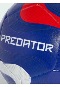 Balón de fútbol azul y rojo con superficie texturizada, que presenta el logo blanco "PREDATOR" y líneas curvas para un mejor agarre. Costura notable visible.