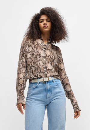Schlangehaut-musterBluse in Beige- und Brauntönen, mit Knopfverschluss und langen Ärmeln, kombiniert mit hellblauen, hoch geschnittenen Jeans und einem weißen Gürtel.