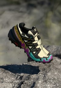 Basket de course en beige et noir avec des accents violets et teal. Dispose d'une semelle texturée, d'un système de laçage et d'un design léger.