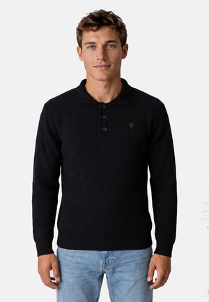 Joven con pelo corto y rizado, vestido con un jersey de polo de punto negro de manga larga y vaqueros azul claro, de pie frente a un fondo liso.