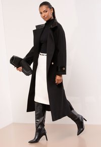 Sort lang frakke med blanke kraveprægninger, kombineret med sort rullekrave, hvid nederdel og sorte knælange teksturerede støvler.