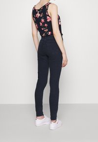 Jeans skinny negros de cintura alta, con un diseño clásico, combinados con una blusa sin mangas de flores. Modelo lleva zapatillas blancas.