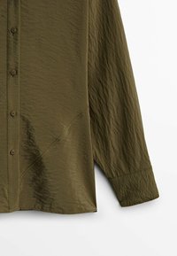 Olivgrön knapp-up skjorta med en texturerad, rynkad tygkvalitet. Har långa ärmar, rak nederkant och diagonala sömmar på sidorna.