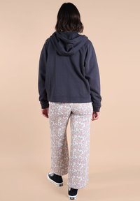 Sweatshirt à capuche gris foncé avec une coupe décontractée, associé à un pantalon large floral clair dans des tons de rose, vert et crème. Des chaussures noires en toile sont visibles.