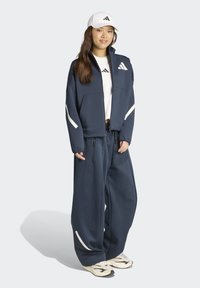 Completo sportivo blu navy con giacca con zip e pantaloni larghi, con dettagli bianchi. Accompagnato da un cappellino bianco con logo.