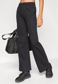 Μαύρο, φαρδύ φούτερ παντελόνι κατασκευασμένο από βαμβακερό μείγμα υφάσματος, με ελαστική μέση, δίπλα σε μια μαύρη αθλητική τσάντα με υφή.