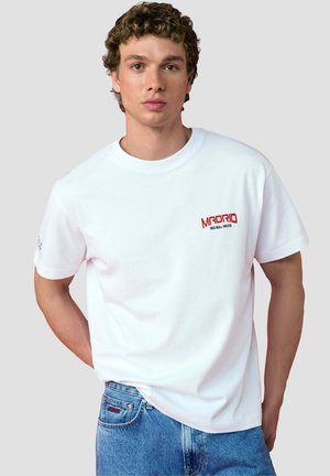 Jeune homme aux cheveux bouclés portant un t-shirt blanc avec "Madrid Red Bull Racing" en rouge et un jean bleu sur un fond gris uni.