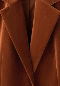 Blazer in velluto marrone con revers a scollo a V e superficie liscia, dal taglio sartoriale e linee pulite, che mette in mostra un colore ricco e profondo.