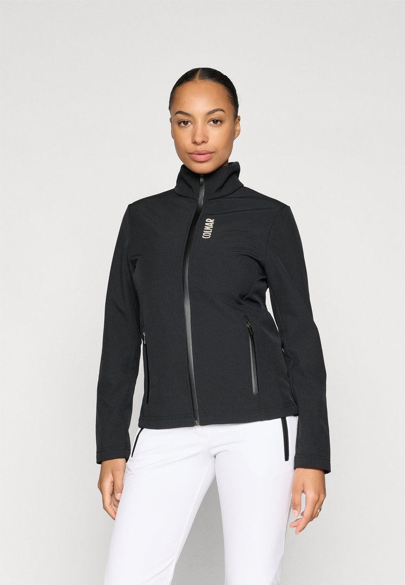 Giacca softshell nera con collo alto, zip frontale e due tasche laterali. Presenta un logo sottile sul lato sinistro del petto. Texture liscia.