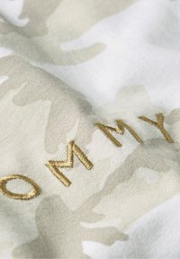 Camouflage-Musterstoff in hellbeige und weiß, mit "TOMMY" in metallisch goldenem Faden gestickt.
