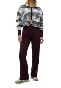 Personne portant une veste courte à carreaux avec de grandes poches, un pantalon bordeaux taille haute et des baskets beige et blanches, debout devant un fond blanc.