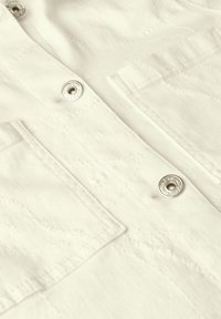 Vaalean beige kangas lähikuvassa, jossa on hopeiset napit, teksturoitu ompelumalli ja tilkkutasku vasemmalla puolella.