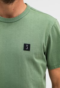 Groene katoenen t-shirt met een ronde hals en korte mouwen. Heeft een zwarte patch met een witte "J" op de linkerborst. Soepele textuur.