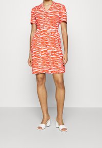 Kvinna som bär en knälång kortärmad klänning i röd och vit mönster med knappar, tillsammans med vita blockklackade sandaler med öppet tå.