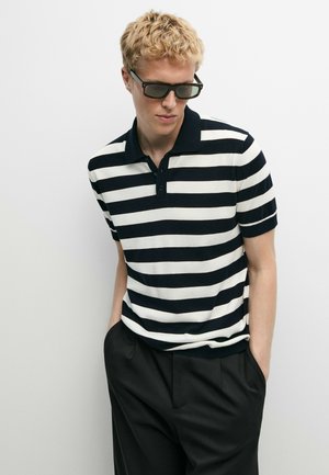 Blonder Mann mit dunkler Sonnenbrille, einem schwarz-weiß horizontal gestreiften Poloshirt und schwarzen Hosen steht mit den Händen in den Taschen.