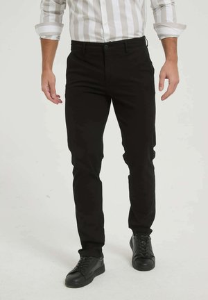 Homme portant un pantalon noir slim, des baskets noires et une chemise blanche aux manches retroussées, debout devant un fond uni.