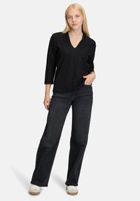 Donna che indossa una maglietta nera a V con maniche tre quarti, jeans larghi scuri, sneakers bianche, in piedi con una mano in tasca.