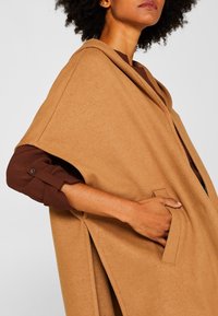 Persona vistiendo un abrigo-capa color camel con un bolsillo lateral, mano dentro del bolsillo, y una camisa de manga larga marrón arremangada debajo.