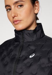 Black puffer jacket featuring a high collar, quilted texture with raised circular patterns, and a zippered front. Small logo on the side.