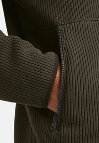 Olivgrüner, gerippter Pullover mit einer seitlichen Reißverschlusstasche. Der Stoff hat eine weiche Textur mit kontrastierenden schwarzen Reißverschlussbeschlägen.